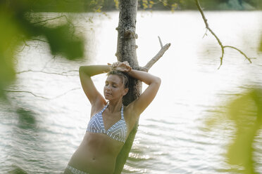 Relaxed woman wearing a bikini leaning against tree trunk at a lake - KNSF05180