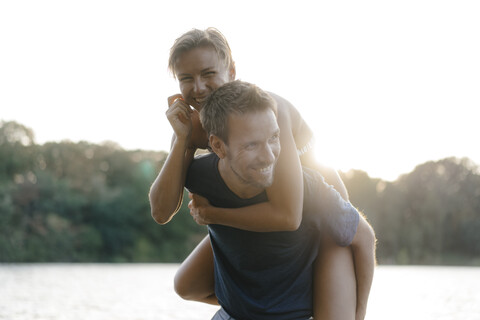 Glücklicher Mann trägt Freundin huckepack an einem See, lizenzfreies Stockfoto