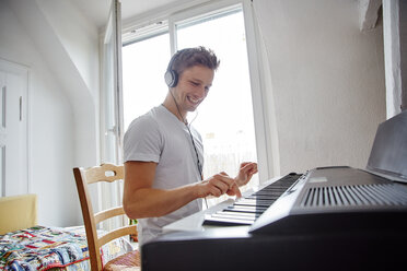 Lächelnder junger Mann, der zu Hause Kopfhörer trägt und Digitalpiano spielt - RHF02267