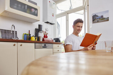 Lächelnder junger Mann sitzt am Tisch zu Hause und liest ein Buch - RHF02283