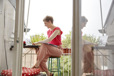 Glückliche Frau sitzt mit Laptop und Landkarte auf dem heimischen Balkon - RHF02292