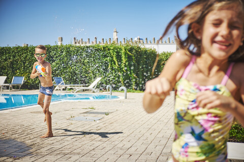 Junge mit Wasserpistole bespritzt Mädchen am Beckenrand, lizenzfreies Stockfoto