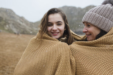 Portrait carefree mother and daughter wrapped in a blanket on snowy winter beach - HOXF04003