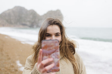 Teenage girl with camera phone taking selfie on winter ocean beach - HOXF04024