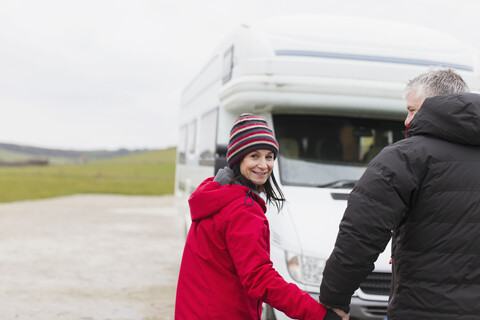 Porträt eines lächelnden Paares in warmer Kleidung, das sich vor einem Wohnmobil an den Händen hält, lizenzfreies Stockfoto