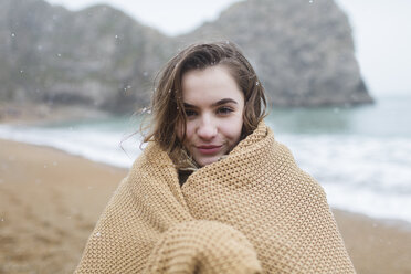 Portrait smiling teenage girl wrapped in blanket on snowy winter beach - HOXF04030