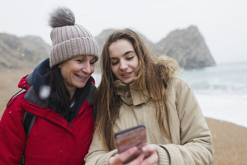 Mutter und Tochter in warmer Kleidung mit Smartphone am verschneiten Winterstrand - HOXF04031