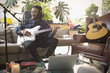 Young male musician recording music, playing guitar and singing into microphone in apartment - HOXF04066