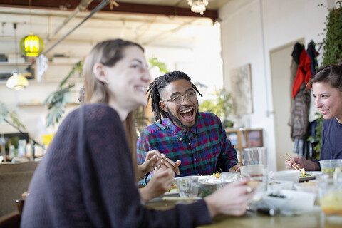 Glückliche junge erwachsene Mitbewohner essen Essen zum Mitnehmen am Küchentisch in der Wohnung, lizenzfreies Stockfoto