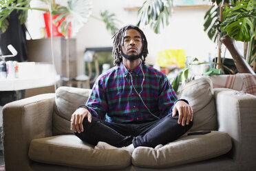 Gelassener junger Mann meditiert mit Kopfhörern auf dem Sofa einer Wohnung - HOXF04090