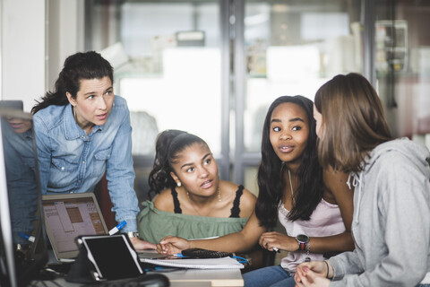 Reife Lehrerin betrachtet selbstbewusste Schülerinnen, die über einen Laptop im Computerraum diskutieren, lizenzfreies Stockfoto