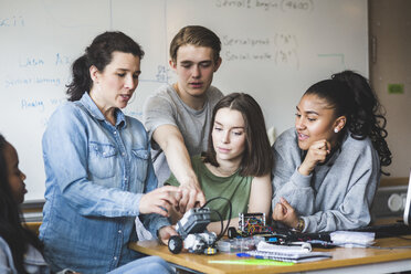 Älterer Lehrer, der einen Roboter untersucht, während er inmitten von Schülern vor der Tafel im Klassenzimmer einer High School sitzt - MASF09291