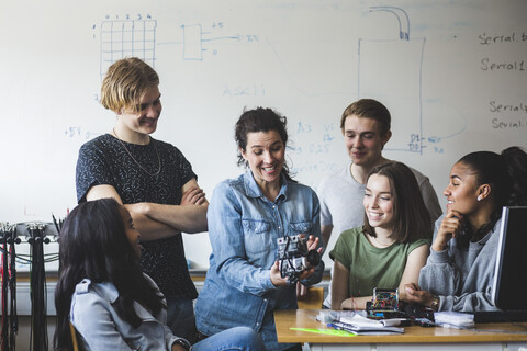 Überrumpelter Lehrer untersucht Roboter, während er inmitten von Schülern vor der Tafel im Klassenzimmer einer High School sitzt, lizenzfreies Stockfoto