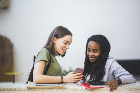 Lächelnde multiethnische Studentinnen, die sich ein Smartphone teilen, während sie im Korridor einer High School stehen, lizenzfreies Stockfoto