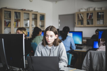 Confident female student using laptop at desk against teacher and friends sitting in computer lab - MASF09315