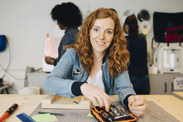 Portrait of smiling redhead female technician sitting at workbench while colleagues working in creative office - MASF09325