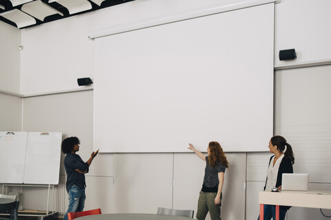 Selbstbewusste multiethnische Techniker halten eine Präsentation über eine leere Projektionsfläche im Kreativbüro, lizenzfreies Stockfoto