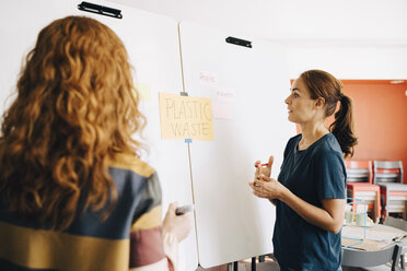 Confident female colleagues discussing plastic management project over whiteboard at creative office - MASF09367