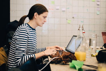 Seitenansicht einer jungen Geschäftsfrau, die in einem kleinen Büro am Schreibtisch sitzend einen Laptop verschlüsselt - MASF09377