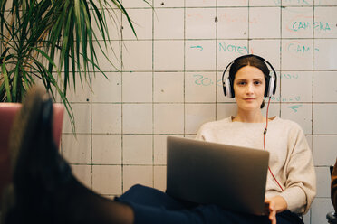 Porträt einer selbstbewussten jungen Computer-Hackerin, die über Kopfhörer zuhört, während sie mit ihrem Laptop an einer Kachelwand sitzt - MASF09386