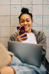 Smiling young female hacker using smart phone while sitting with laptop against tile wall at small creative office - MASF09392