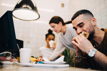 Junger männlicher Hacker, der ein Sandwich isst, während seine weiblichen Kollegen während einer Besprechung in einem kleinen Büro arbeiten - MASF09403
