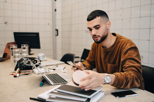 Junger männlicher Hacker, der ein Smartphone benutzt, während er ein Gerät am Schreibtisch in einem kleinen kreativen Büro hält - MASF09426