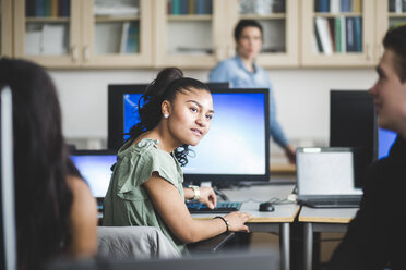 Confident female high school student looking at friend sitting in computer lab - MASF09431