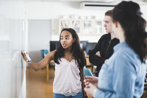 Confident girl explaining from whiteboard to teacher and friend in classroom stock photo