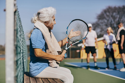 Ältere Frau hält einen Tennisschläger, während sie auf einer Bank am Tennisplatz sitzt, lizenzfreies Stockfoto