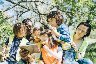 Multi-ethnic students and teachers sharing digital tablet while sitting against trees in playground - MASF09527