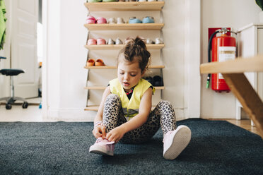 Girl sitting on carpet wearing shoes against rack in cloakroom at child care - MASF09531