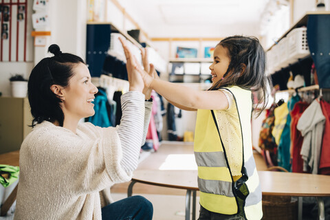 Seitenansicht einer glücklichen Lehrerin und eines Mädchens, die sich in der Garderobe des Kindergartens abklatschen, lizenzfreies Stockfoto