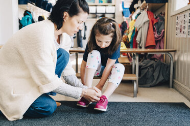 Seitenansicht einer Lehrerin, die einem Mädchen in der Garderobe der Vorschule hilft, Schuhe anzuziehen - MASF09542