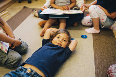 Hochformatiges Porträt eines Jungen, der auf einem Teppich inmitten von Freunden im Klassenzimmer liegt - MASF09547