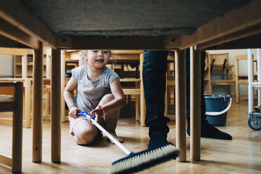 Full length of girl cleaning hardwood floor below table by teacher standing in classroom - MASF09550