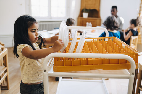 Seitenansicht eines Mädchens, das einen Teller in eine Kiste auf einem Wagen im Klassenzimmer stellt, lizenzfreies Stockfoto