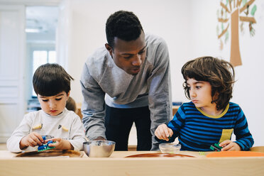 Männlicher Lehrer betrachtet Schüler, die mit Spielzeug am Tisch in einem Kinderbetreuungsraum spielen - MASF09582