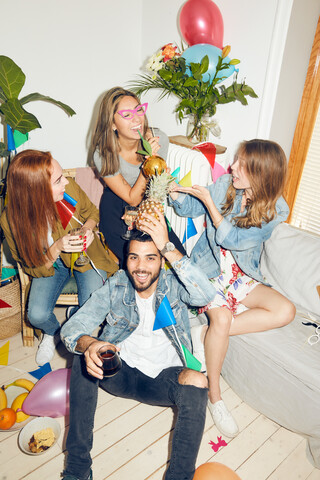 Porträt eines lächelnden jungen Mannes, der eine Ananas in der Hand hält, während er gegen weibliche Freunde sitzt, die eine Party zu Hause genießen, lizenzfreies Stockfoto