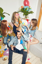 Porträt eines lächelnden jungen Mannes, der eine Ananas in der Hand hält, während er gegen weibliche Freunde sitzt, die eine Party zu Hause genießen - MASF09605