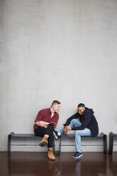 Volle Länge der glücklichen männlichen Studenten mit Technologien sitzen auf Bank gegen die Wand in der Universität - MASF09633
