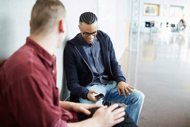 Junger Mann benutzt sein Smartphone, während er mit einem Freund auf einer Bank im Korridor der Universität sitzt - MASF09636