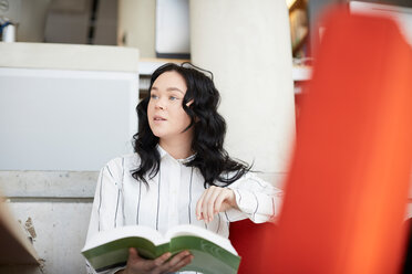 Junge Frau mit Buch, die wegschaut, während sie in der Cafeteria einer Universität sitzt - MASF09663