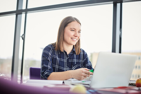 Lächelnde junge Studentin, die einen Laptop benutzt, während sie am Tisch in der Universitätscafeteria lernt, lizenzfreies Stockfoto