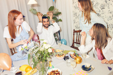 Playful man holding pineapples while sitting amidst female friends at home during dinner party - MASF09696