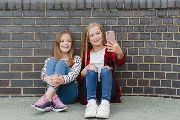 Porträt von zwei lächelnden Mädchen, die vor einer Backsteinmauer sitzen und ein Selfie mit ihrem Smartphone machen - NMSF00284
