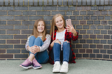 Portrait of two smiling girls sitting in front of brick wall taking selfie with smartphone - NMSF00284