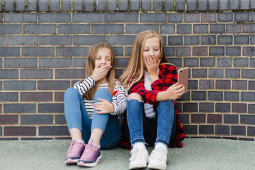 Porträt von zwei Mädchen, die vor einer Backsteinmauer sitzen und ein Selfie mit ihrem Smartphone machen - NMSF00285