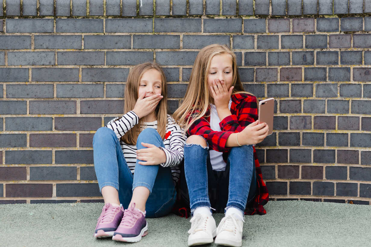 Portrait of two girls sitting in front of brick wall taking selfie