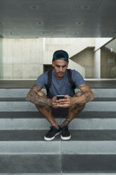 Tattooed young man sitting on stairs using smartphone - JPTF00050